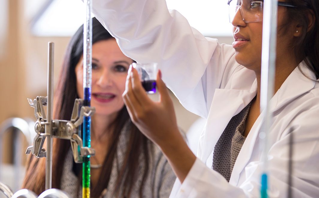 Dr. Michelle Hogue looks on as ULethbridge student in lab coat holds beaker of solution