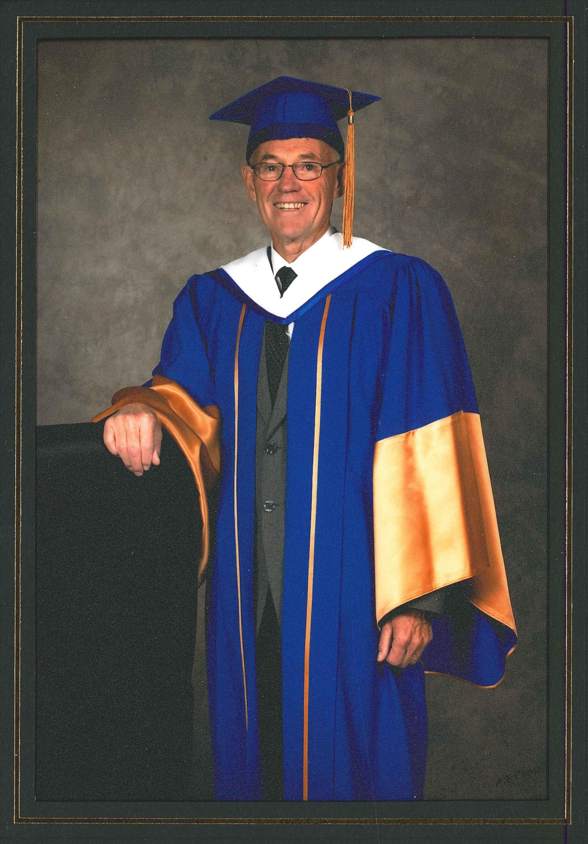 An older man with glasses wears a blue and gold university robe and cap, with his right hand resting on a chair.