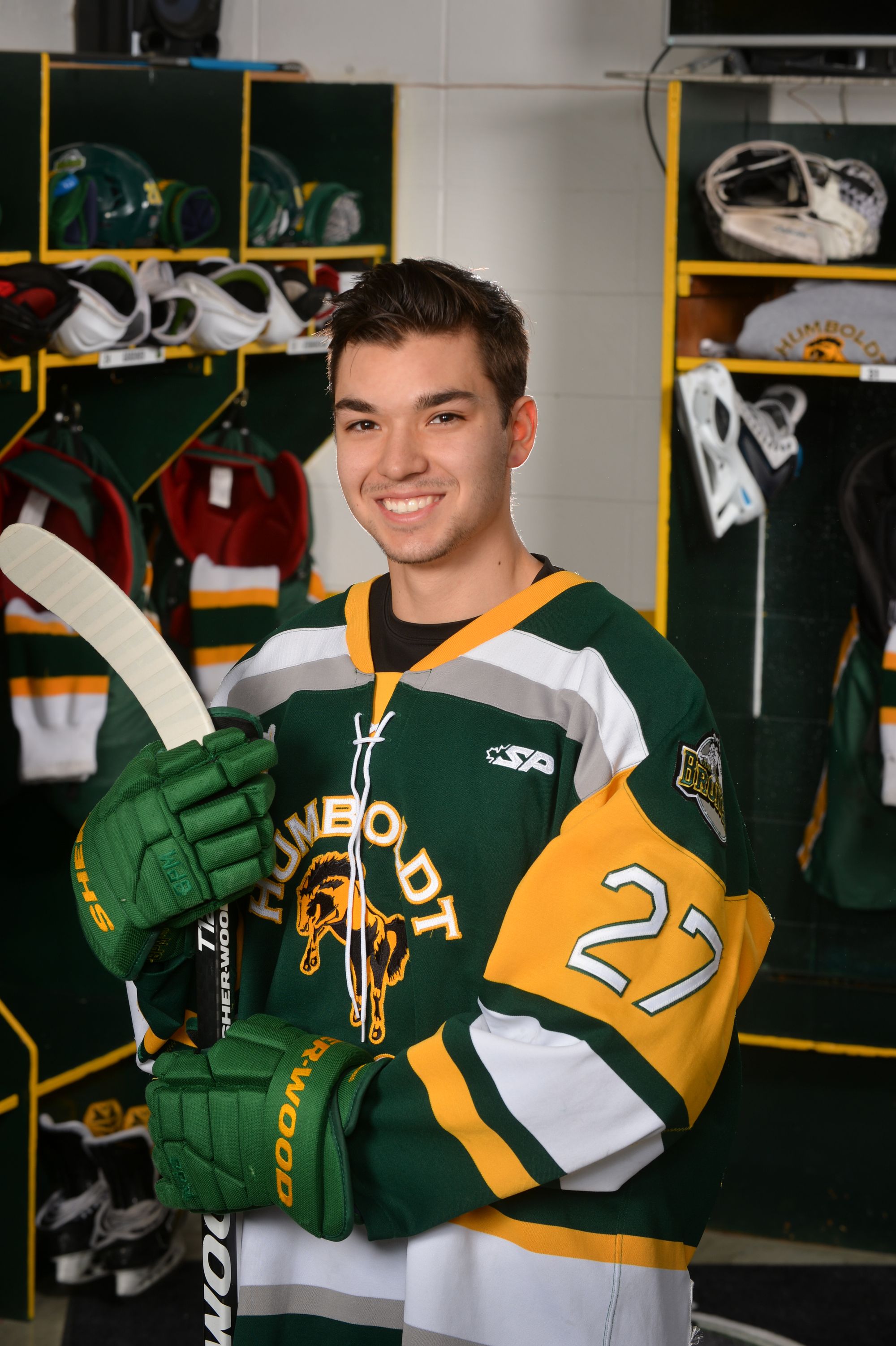 A young man wears a hockey jersey and holds a hockey stick. The jersey is yellow, green, and white with the number 27.