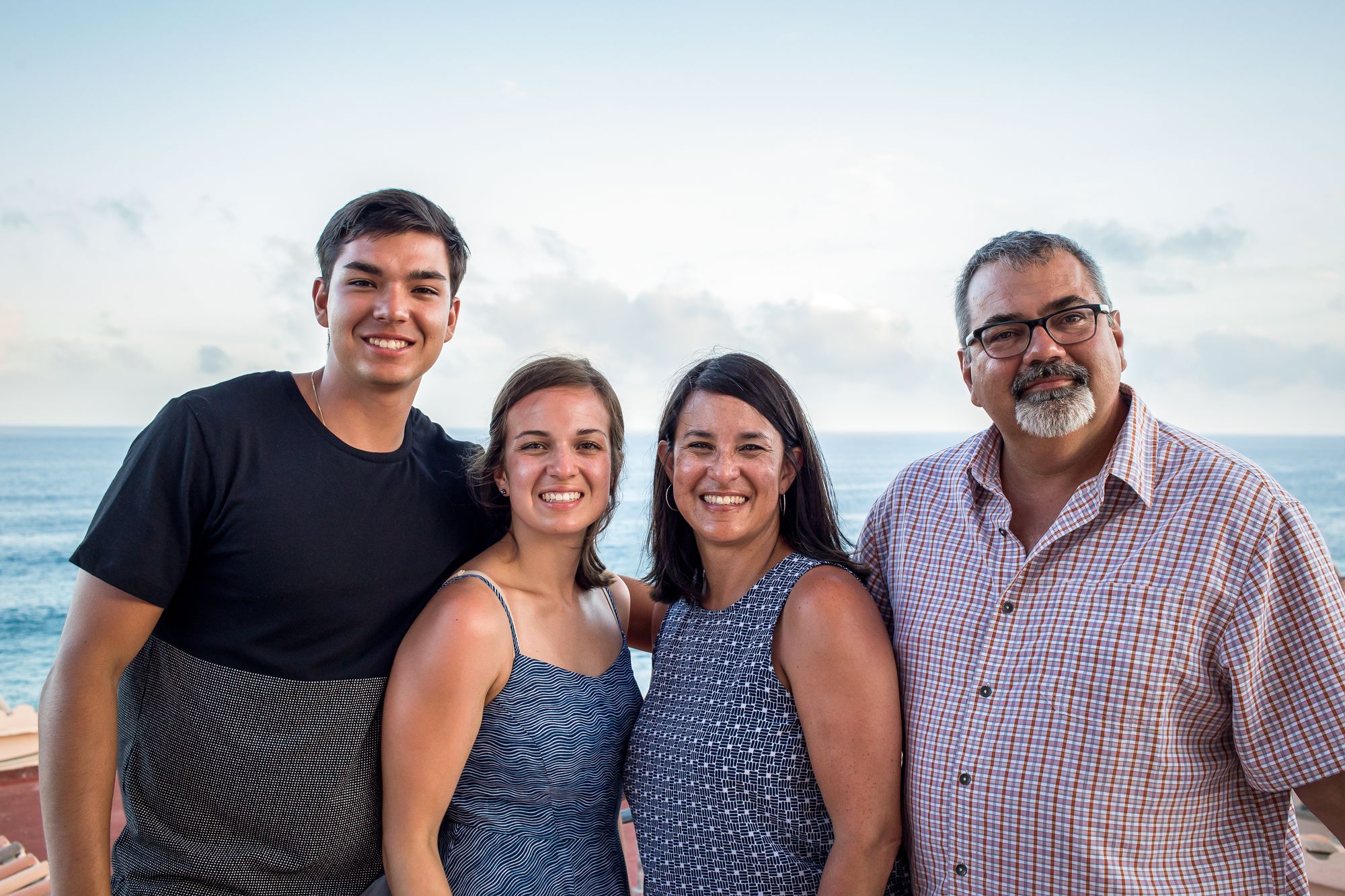A family of four stand smiling in front of an ocean. A young man wearing a navy shirt, a young woman wearing a blue dress, a woman wearing a blue tank top, a man with glasses and a grey beard wearing a red and blue plaid shirt.