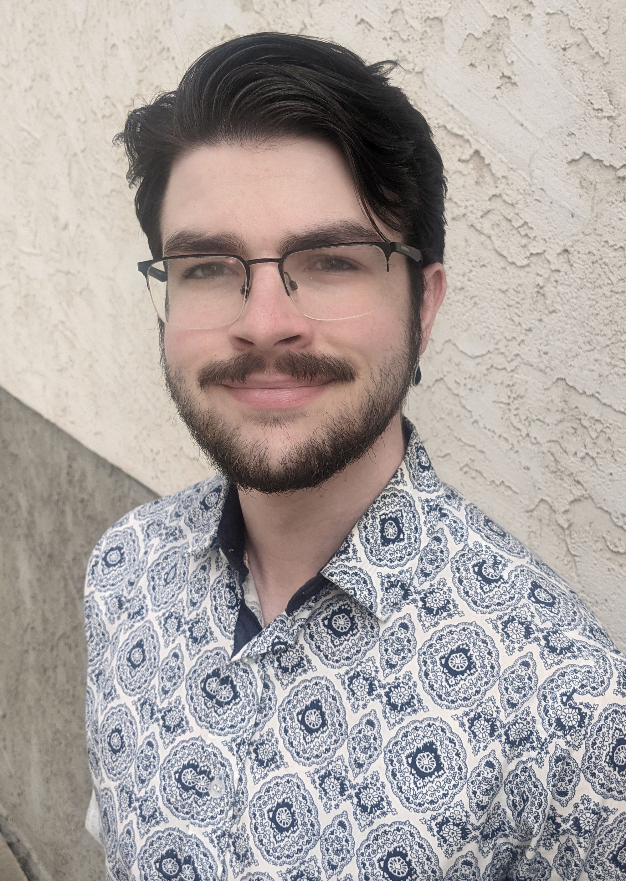 A portrait of a young man with glasses, short black hair, and a beard. He is wearing a blue paisley button up shirt.