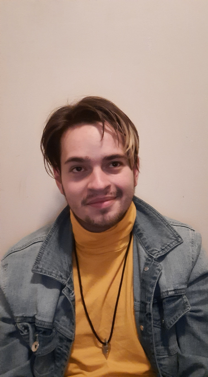 Portrait of a young man with brown hair and facial hair, wearing a jean jacket and yellow turtleneck and a necklace.