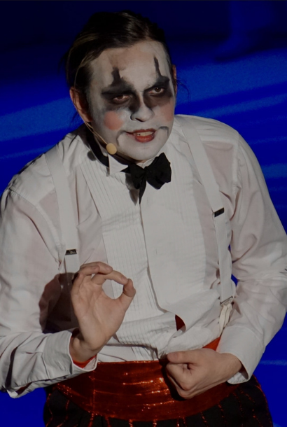 A male actor in a play, wearing clown makeup with a white shirt, suspenders, a bowtie and red pants.