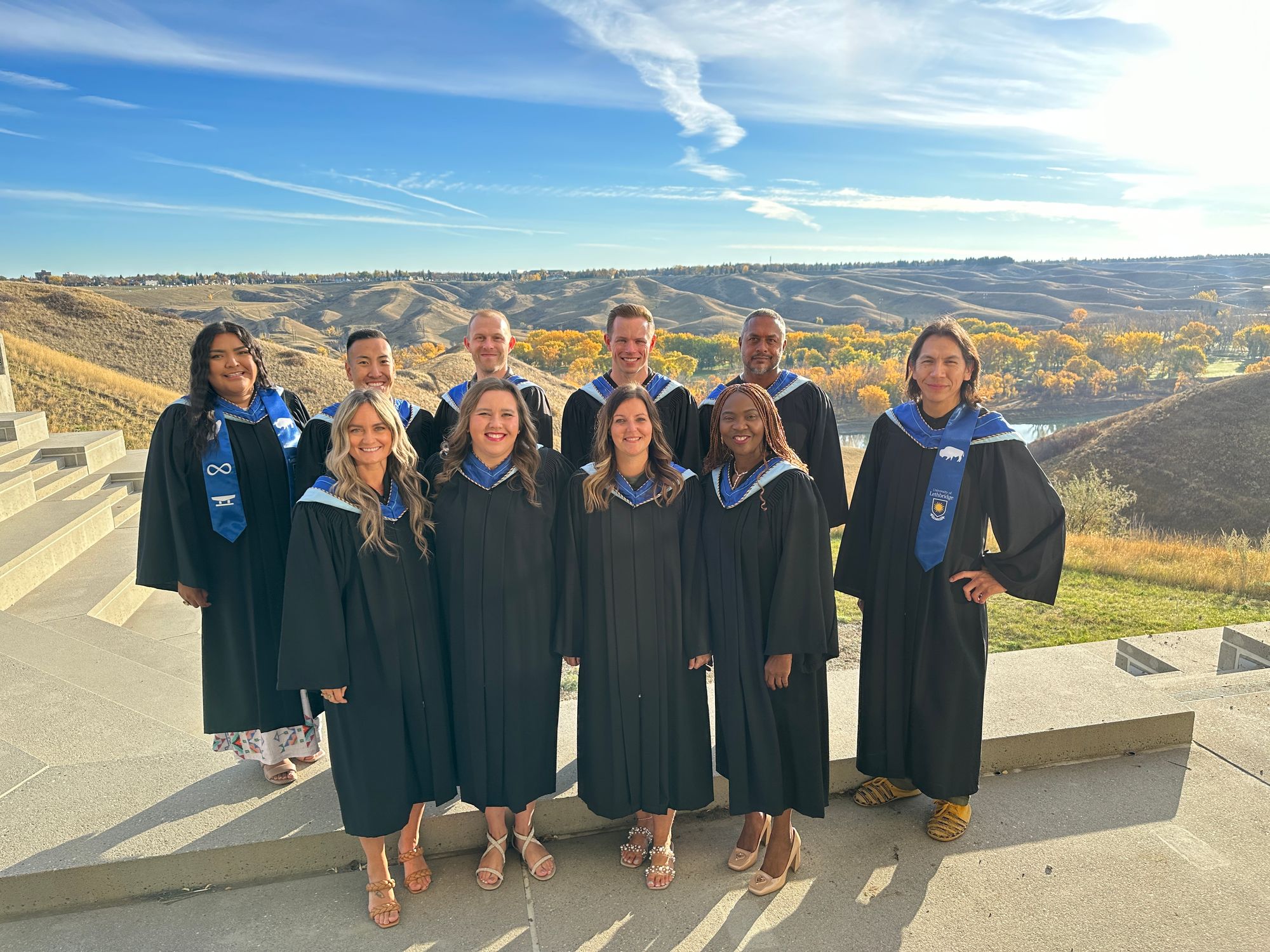 Group of 10 ULethbridge Master of Education graduates in black regalia with blue hoods with Oldman river valley in background.