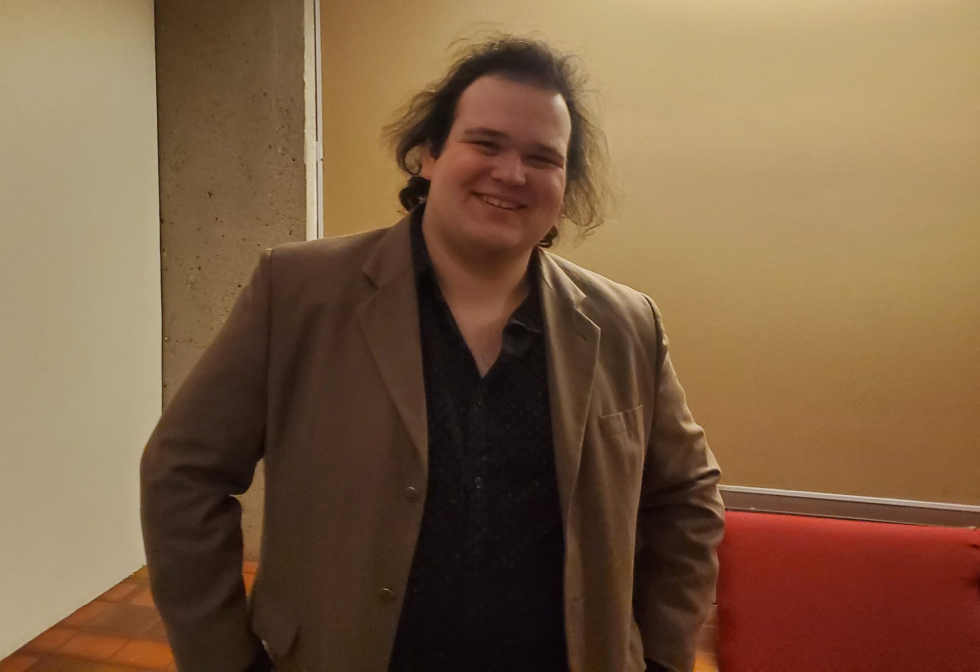 A person with shoulder-length dark hair, wearing a tan blazer and dark button-up shirt.